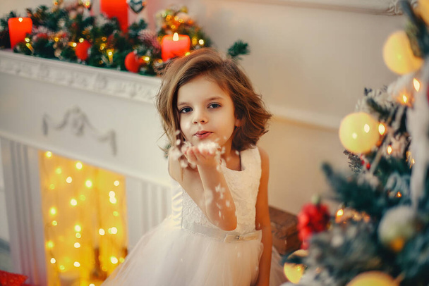 Cute little girl in white holiday dress standing in brightly decorated room, near Christmas tree, full of decoration, blowing snow from hand. New Year concept  - Φωτογραφία, εικόνα