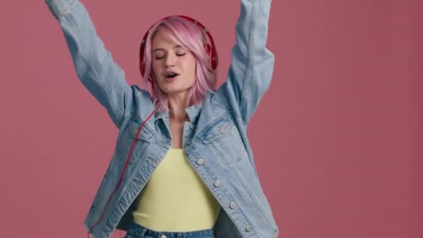 Προφίλ του hipster έφηβος όμορφο μοντέλο κορίτσι της μόδας φορούν μοντέρνα ακουστικά απολαμβάνοντας χορό, ακούγοντας νέα δροσερή μουσική μίξη σταθεί σε ροζ φόντο στούντιο σε μοντέρνα τζιν σακάκι κίτρινο κορυφή 4K - Πλάνα, βίντεο