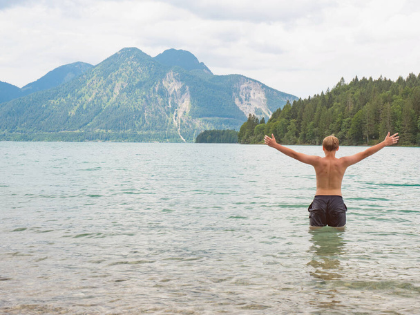 10代の少年は水の奥深くに腰を立て、腕を上げて敬礼する。アルパイン湖での水泳,ヘルツォグスタンドのピーク,バイエルン州北部. - 写真・画像
