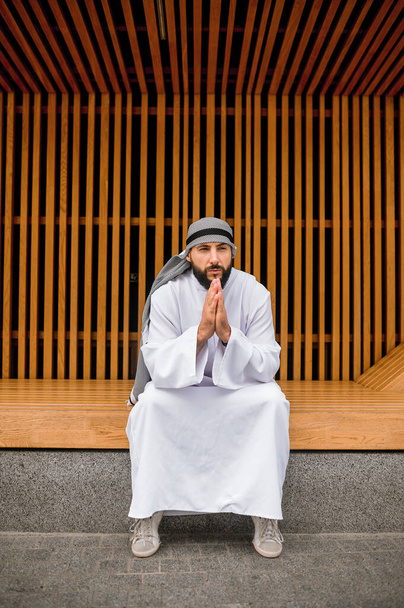Dinleniyor. Ortadoğulu genç adam geleneksel giysiler içinde tahta bir bankta oturuyor. - Fotoğraf, Görsel