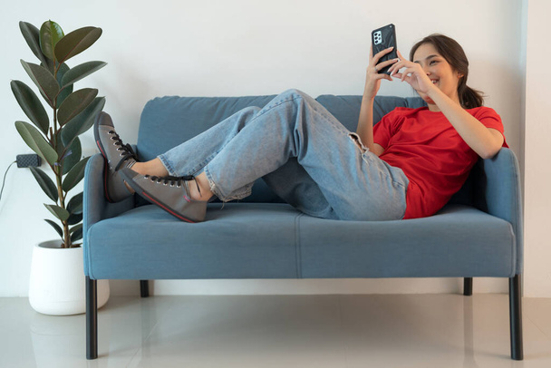 Una bella donna con una camicia rossa sdraiata sul divano che gioca con il suo telefono Naviga in Internet in un umore rilassato e felice. - Foto, immagini