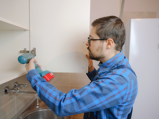 young man in blue work suit using electric screwdriver when assembling cupboard in kitchen. Home renovation concept. - Zdjęcie, obraz
