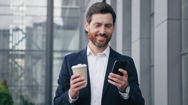 Successful smiling handsome businessman walking down street holding paper cup with coffee looks at smartphone screen browsing social network reading good news on mobile phone communicates on internet - Footage, Video