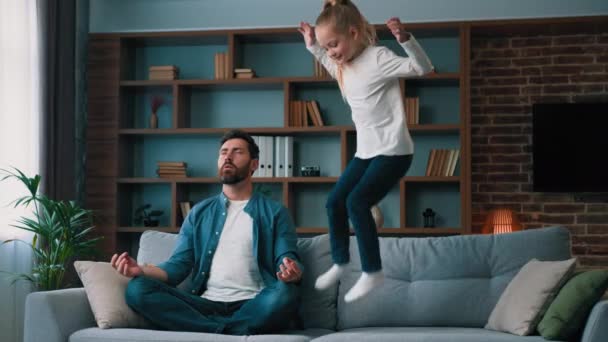 Rauhallinen rauhallinen mies isä istuu sohvalla lootuksessa aiheuttaa joogaa käytännössä terveyden virkistys kotona rauhallinen isä huomiotta aktiivinen energinen tuhma lapsi tyttö lapsi lapsi hyppää pelaa häiritä vanhemman meditaatio - Materiaali, video