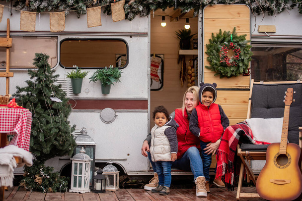 Caucasian mother plays having fun with African American children on the doorstep Trailer house. Young woman hugs kisses son, daughter. The family laughs. Christmas decorations, travel for the holidays - Фото, зображення