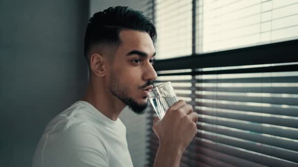 Bezárás beltéri elmélkedő elmélkedő spanyol indián latin szakállas srác millenniumi férfi néz ablak otthon úgy gondolja, álom van törés italok üveg hidratáló víz egyensúly ellátás - Felvétel, videó
