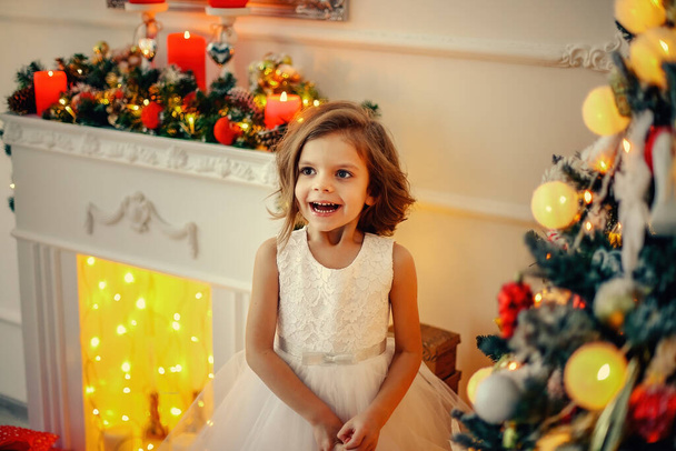 Surprised cute little girl in white dress standing in brightly decorated room near Christmas tree full of decoration. New Year concept  - 写真・画像