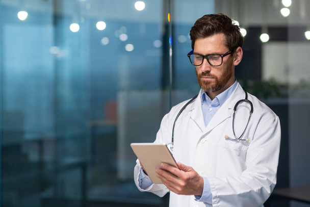 Ein seriöser und nachdenklicher Arzt liest medizinische Informationen von einem Tablet-Computer, ein Mann im Arztkittel bedient sich eines Tablets, konzentriert sich, tippt Nachrichten ein und überprüft die Patientenakte - Foto, Bild