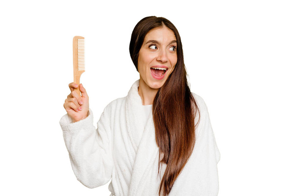 Mulher caucasiana jovem segurando um pente de cabelo isolado olha para o lado sorridente, alegre e agradável. - Foto, Imagem