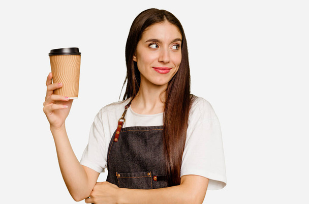 Young barista woman holding a takeaway coffee isolated looks aside smiling, cheerful and pleasant. - Photo, Image