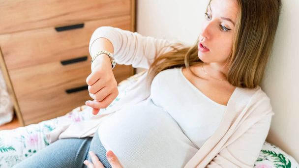 Беременные схватки. Беременная женщина смотрит на часы, держит животик. Время родов, схватки боль. Беременность, медицина - Фото, изображение