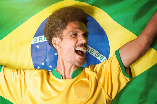 Abanico de fútbol brasileño - Hombre negro sosteniendo la bandera de Brasil detrás de la celebración - Foto, imagen