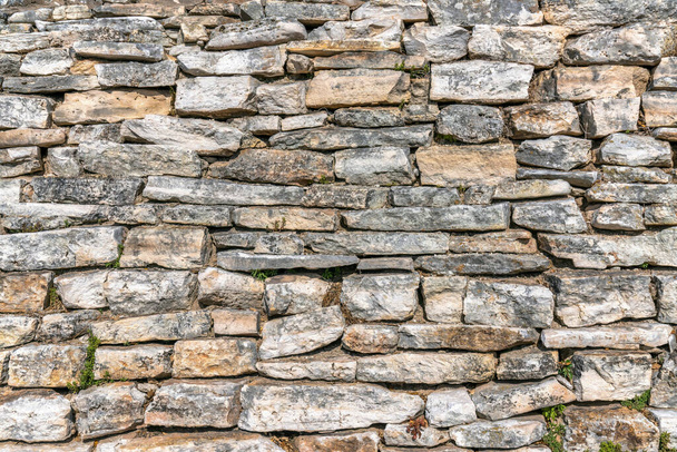 Oude bakstenen muur met kleine groene planten die groeien tussen de onregelmatige blokken. Close-up zicht op een stenen oppervlak van een gebouw of muur met een ruwe textuur en ruw patroon. - Foto, afbeelding