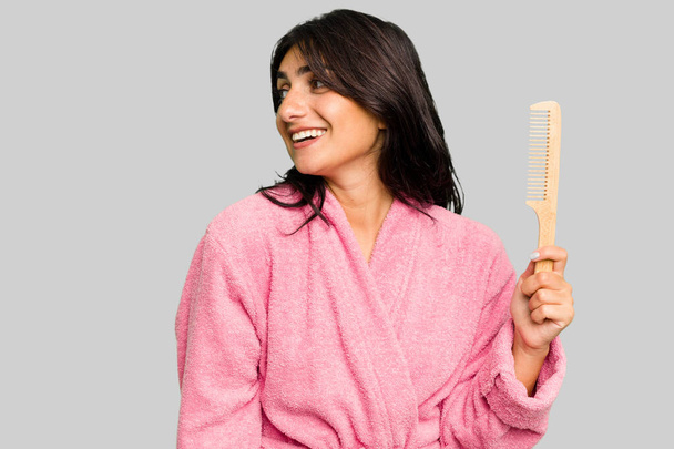 Mulher jovem em um roupão de banho segurando um pente de cabelo isolado olha para o lado sorridente, alegre e agradável. - Foto, Imagem