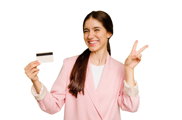 Mujer caucásica de negocios sosteniendo una tarjeta de crédito aislada alegre y despreocupada mostrando un símbolo de paz con los dedos. - Foto, imagen