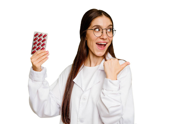 Young pharmacist woman holding a tablet of pills isolated points with thumb finger away, laughing and carefree. - Φωτογραφία, εικόνα