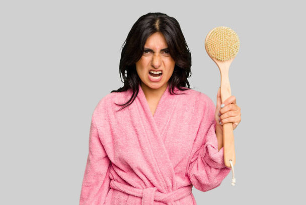 Young Indian woman in a bathrobe holding a back scratcher isolated screaming very angry and aggressive. - Photo, Image