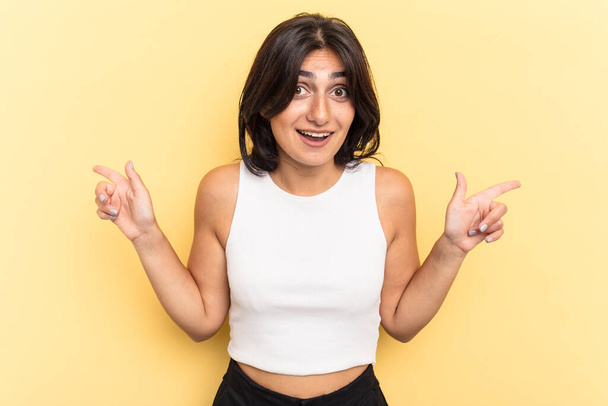Young Indian woman isolated on yellow background pointing to different copy spaces, choosing one of them, showing with finger. - Photo, Image