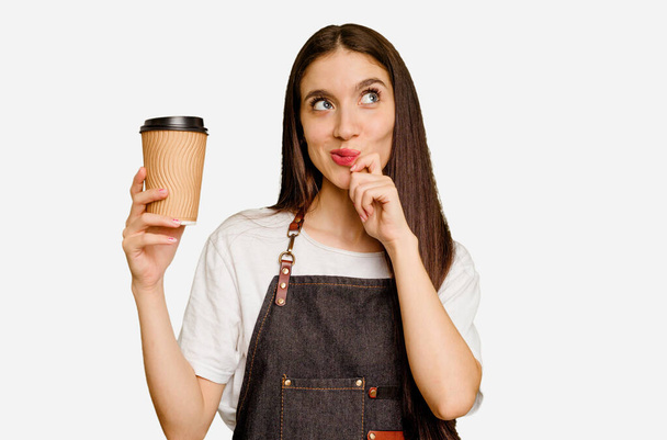 Молодая бариста держит кофе на вынос изолированный глядя в сторону с сомнительным и скептическим выражением лица. - Фото, изображение