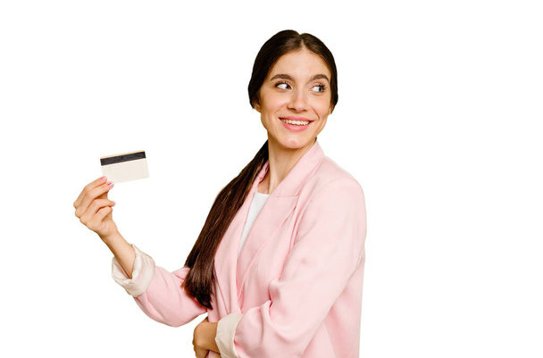 Mulher caucasiana de negócios segurando um cartão de crédito isolado olha para o lado sorridente, alegre e agradável. - Foto, Imagem