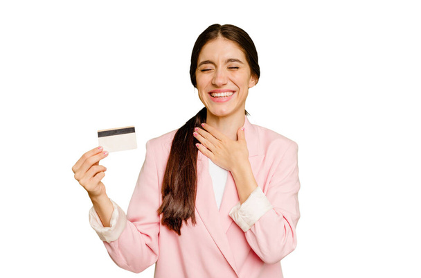 Mujer caucásica de negocios sosteniendo una tarjeta de crédito aislada se ríe en voz alta manteniendo la mano en el pecho. - Foto, imagen