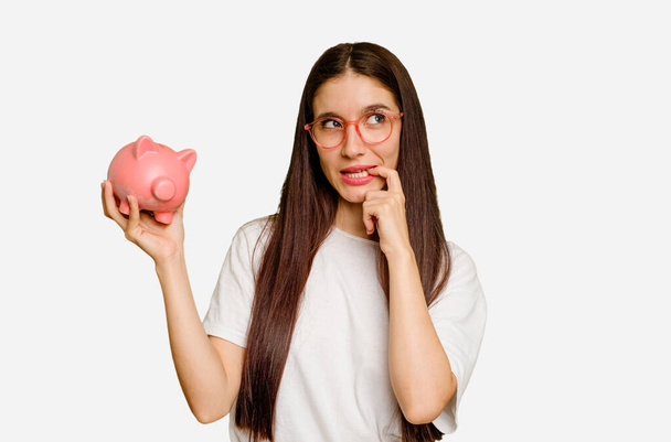 Young caucasian woman holding a piggy bank isolated relaxed thinking about something looking at a copy space. - Photo, Image