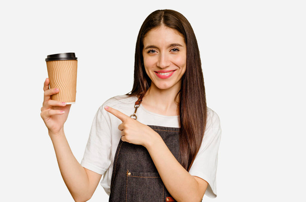 Jonge barista vrouw houdt een afhaalkoffie geïsoleerd glimlachend en wijzend opzij, toont iets op lege ruimte. - Foto, afbeelding