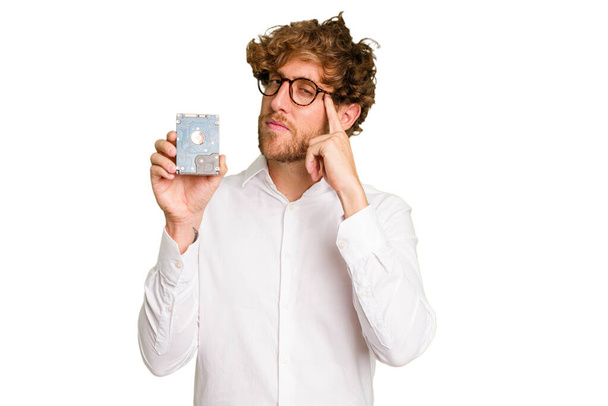 Joven hombre informático caucásico sosteniendo un disco duro aislado sobre fondo blanco apuntando templo con el dedo, pensando, centrado en una tarea. - Foto, imagen