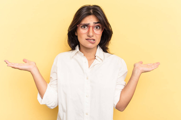 Young Indian woman isolated on yellow background confused and doubtful shrugging shoulders to hold a copy space. - Foto, immagini