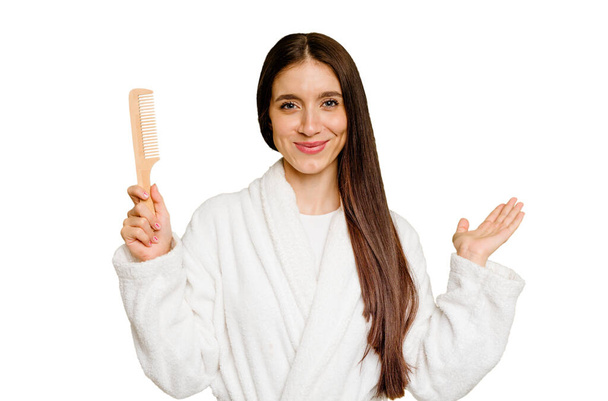 Mulher branca jovem segurando um pente de cabelo isolado mostrando um espaço de cópia em uma palma e segurando outra mão na cintura. - Foto, Imagem