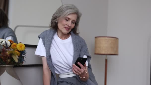μια γυναίκα σε ηλικία συνταξιοδότησης με κομψά ρούχα και στο σπίτι διαβάζει μηνύματα στην οθόνη ενός κινητού τηλεφώνου. - Πλάνα, βίντεο