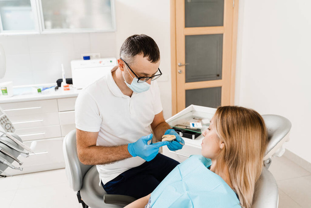 Οδοντίατρος δείχνει γύψο των δοντιών της ασθενούς γυναίκας πριν από την οδοντική εμφύτευση. Διαδικασία δημιουργίας οδοντιατρικών προθέσεων, στεφανών και αλλοδαπών - Φωτογραφία, εικόνα