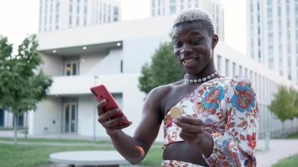 Fiatal fekete afrikai transznemű férfi bitcoinnal a kezében és egy mobiltelefonnal izgatott a kriptovaluta befektetése miatt. - Felvétel, videó