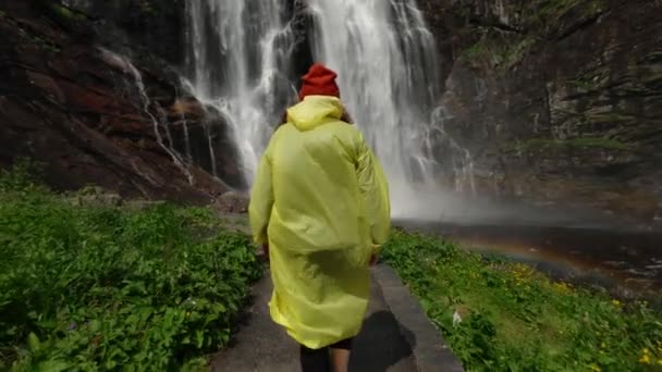 Młoda dziewczyna wędrowiec w żółtym płaszczu idzie do wodospadu w górach, podnosi ręce do góry, cieszy się przyrodą i życiem. Podróżowanie w górach, przygoda w podróży. Koncepcja stylu życia. - Materiał filmowy, wideo