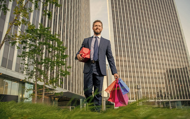 веселый харизматичный зрелый бизнесмен в костюме держать пакеты и подарочные коробки за пределами офиса, случай. - Фото, изображение
