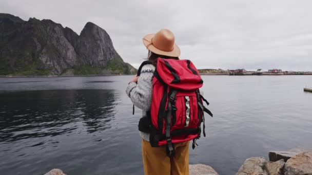 De reiziger gaat naar de houten pier. gekleed in een trui en een gebreide muts en een rode rugzak. Achteraanzicht. Lofoten-eilanden Noorwegen. - Video