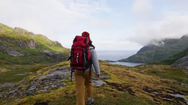 A girl with a big backpack hikes through the highlands of Norway. The trail is barely marked. Fresh green grass. Taller mountains in the back covered partially with snow. Wilderness and freedom - Footage, Video