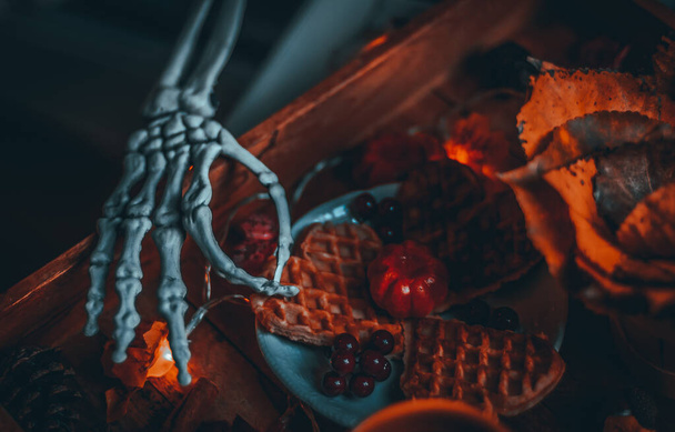 Skelet hand reikt naar wafels in een houten dienblad met pompoen, bessen, kegels, populier bladeren en een brandende bloemenslinger liggend op een nachtkastje, close-up zijaanzicht met scherptediepte. Halloween ontbijt concept. - Foto, afbeelding