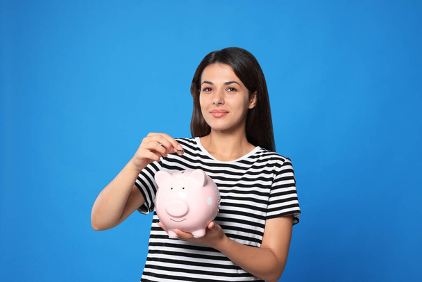 Young woman putting coin into piggy bank on light blue background - 写真・画像