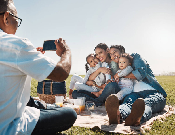 Family, phone and picnic photo with smile, hug and happy together in summer on grass in park. Grandparents, mother or children with smartphone picture, social media and online during break on weekend. - Photo, Image