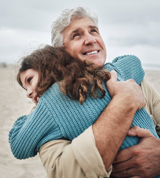Family, children and hug with a girl and grandfather embracing on the beach outdoor during a holiday or vacation. Travel, kids and love with a senior man thankful for his granddaughter by the sea. - Photo, Image