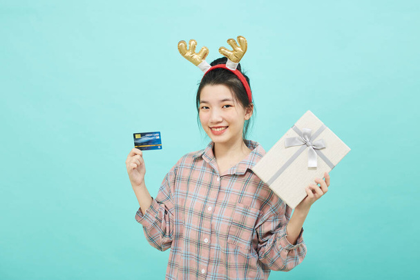 Cheerful cute Asian woman holding a gift box with a credit card on bright blue color background. Christmas and new year sales joy concept - Zdjęcie, obraz