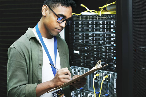 Skilled in and abled in the handling of wires and cables. a young man making notes while working in a server room - Photo, Image