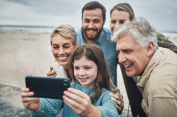 Family, selfie and girl with phone on beach for holiday, vacation or journey together by ocean. Group, mom and dad with child, grandparents and smartphone for photo in winter with smile by sea. - Foto, Imagem