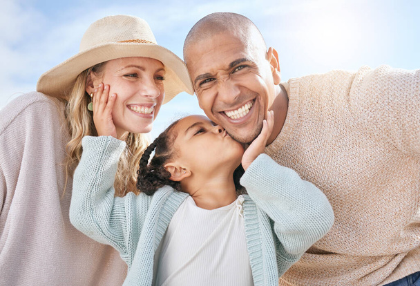 Happy family, mother and father with a kiss from a child on the face in summer outdoors on a weekend. Interracial, mama and girl loves kissing dad with a big smile on cheek on sunny holiday vacation. - Photo, Image