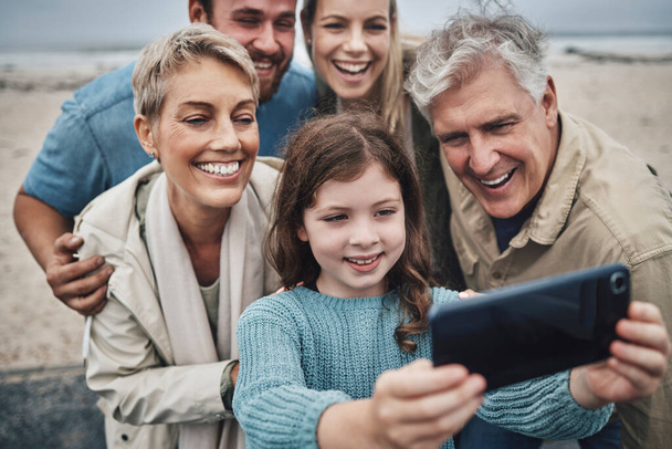 Familie, Mädchen und Handy-Selfie im Strandurlaub, Zeit mit Eltern und Großeltern verbringen. Männer, Frauen und Kinder lächeln in Australien, fröhlicher Spaß bei der Winterwanderung am Meer und Foto auf dem Smartphone. - Foto, Bild