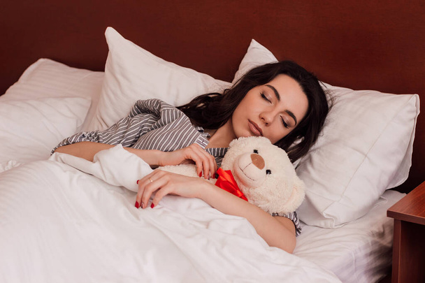 A young beautiful girl sleeps with a teddy bear in a bed in pajamas. High quality photo - 写真・画像
