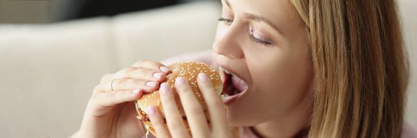 Portrait of young blonde female eating juicy hamburger, bite junk food, fatty lunch, order fast food to home. Unhealthy eating, takeaway, hunger concept - Photo, Image