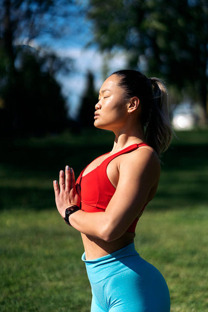 Concentrated asian woman stretching after workout in the park. She has her eyes closed and her hands together. - Fotoğraf, Görsel