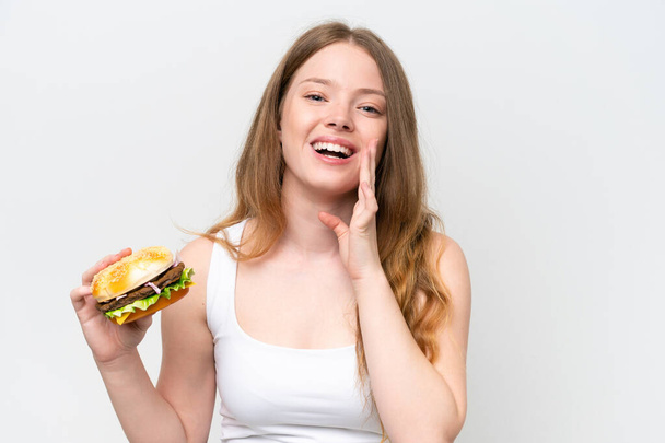 Nuori kaunis nainen tilalla hampurilainen eristetty valkoisella taustalla huutaa suu auki - Valokuva, kuva
